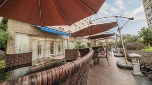 An outdoor seating area with wicker furniture and large umbrellas, a stone building with a blue awning, and a street lamp.