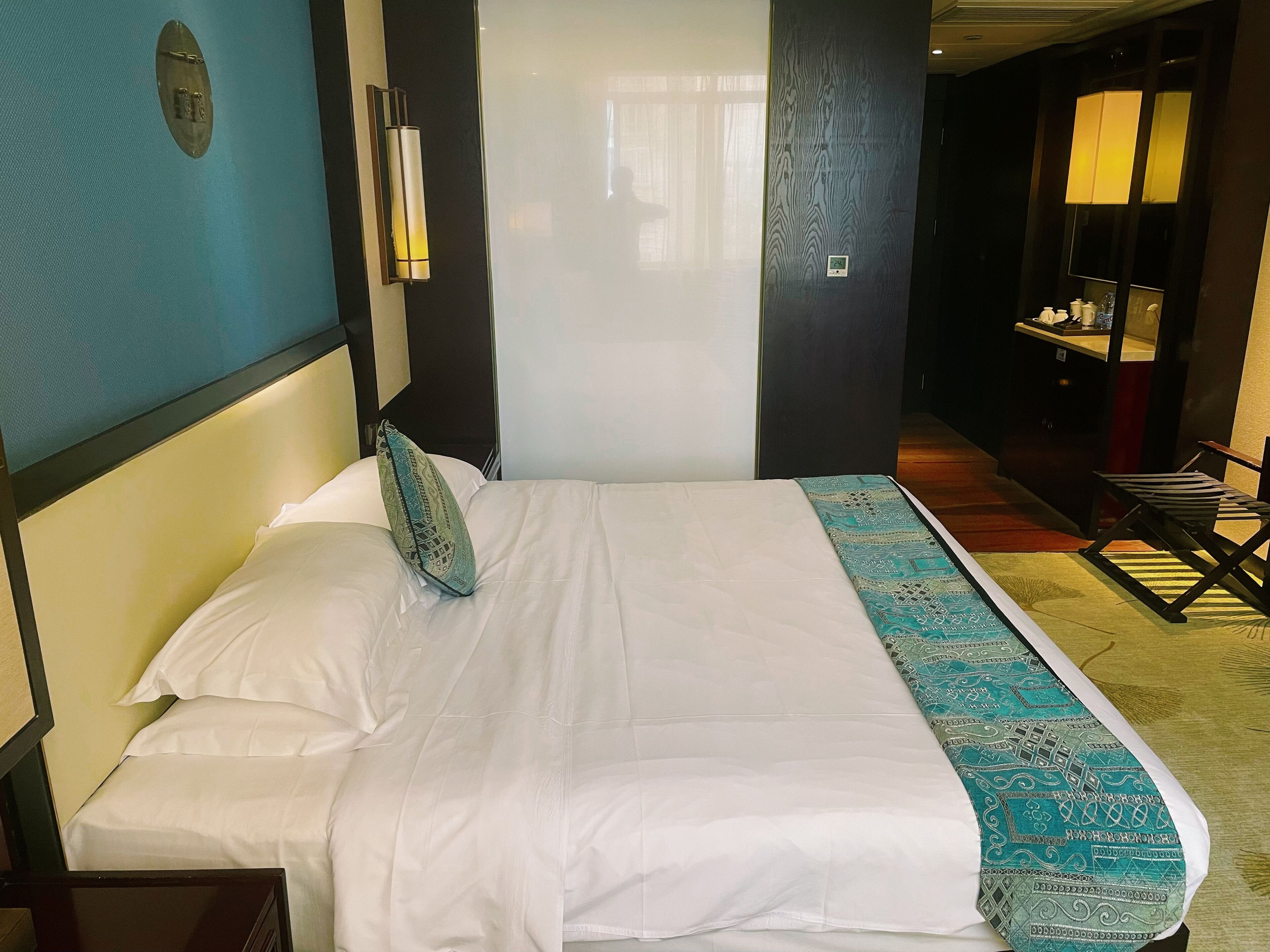 Habitación de hotel con cama, mesita de noche, lámpara y vista al pasillo.