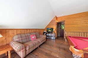 A loft-style room with a wooden sloped ceiling, a plaid sofa with a red cushion, a pool table, and a television in the background.