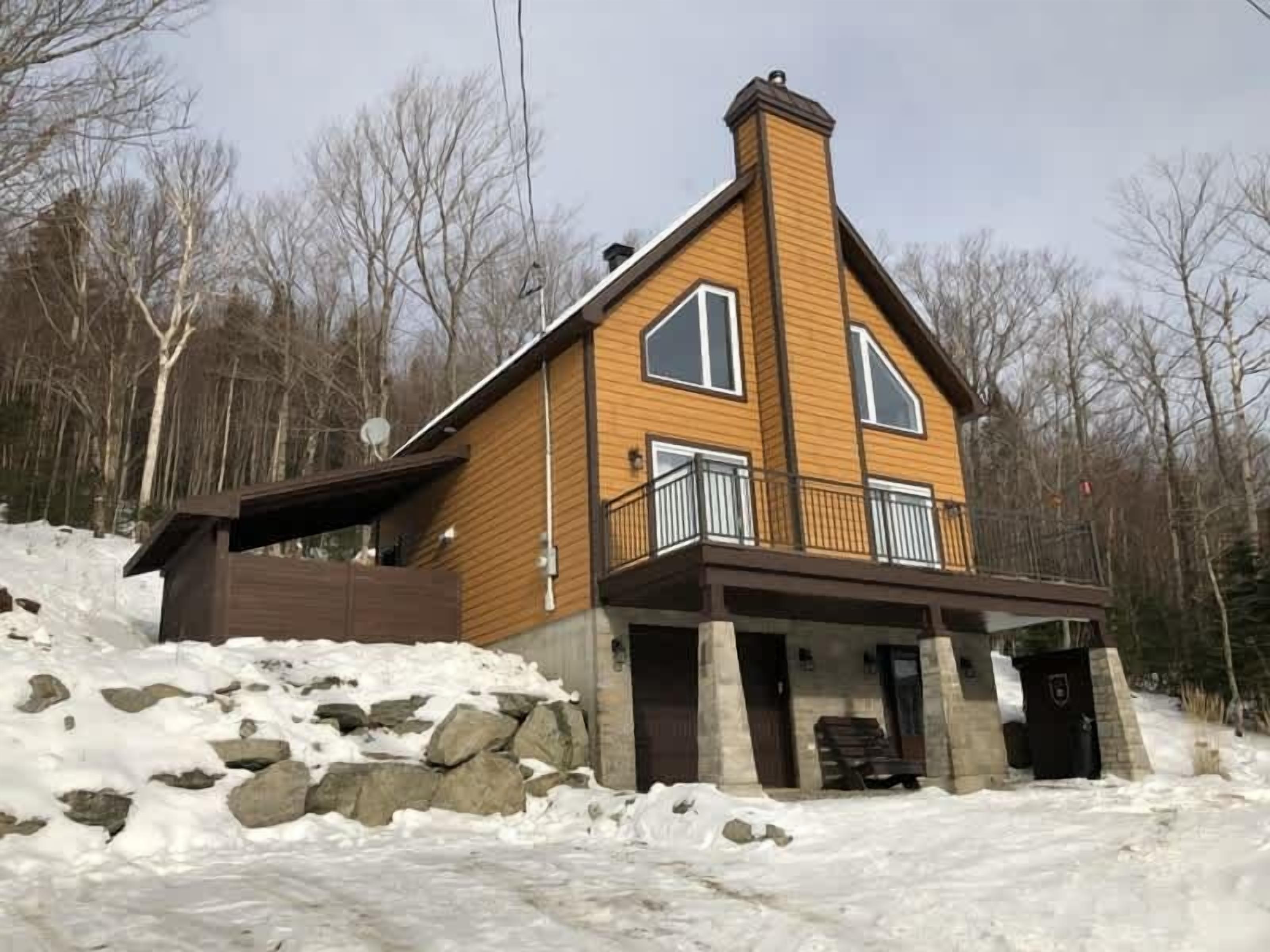 A two-story wooden cabin with a balcony, surrounded by snow and trees.