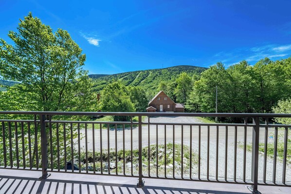 A wooden cabin with a gravel driveway, surrounded by lush greenery and hills in the background.