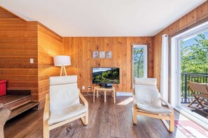Une pièce aux murs lambrés, équipée d’un téléviseur, de deux fauteuils, d’une lampe et d’un balcon offrant une vue panoramique.