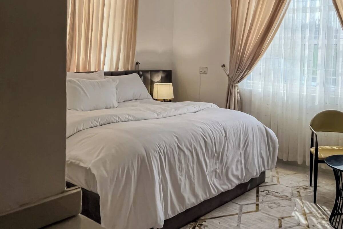 A hotel room with a large bed, a bedside lamp, a chair, and a view of the city through sheer curtains.