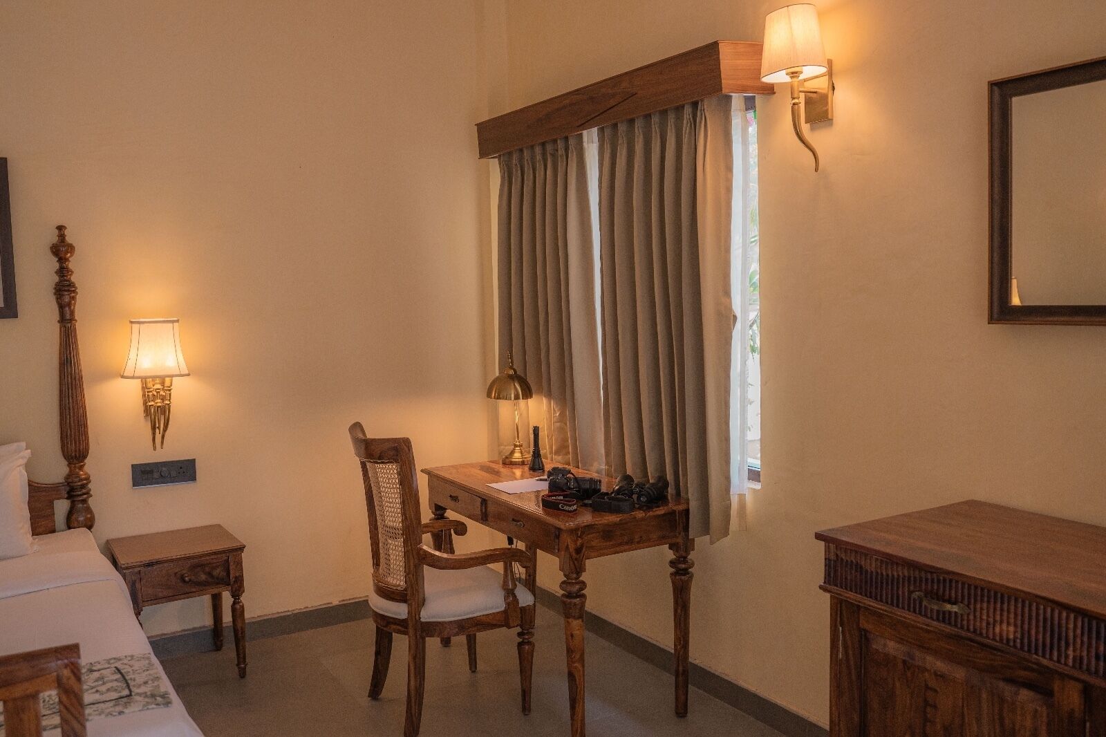 Una habitación con un escritorio de madera, dos lámparas, una silla, una cama y un ventanal con cortinas.