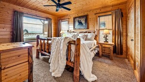 A wooden cabin bedroom with a bed, a desk, a chair, a ceiling fan, and a window with curtains.