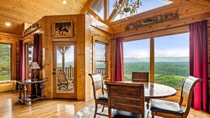 A wooden cabin with a dining area featuring a round table and chairs, a chandelier, and large windows offering a view of the mountains.