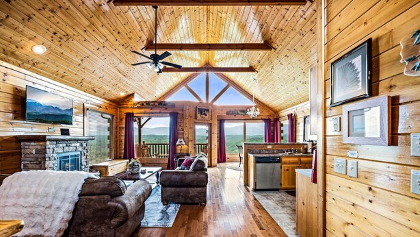 A log cabin interior with a fireplace, wooden floors, a sofa, a dining area, and large windows offering a mountain view.
