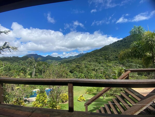 Una terraza de madera con vistas a un paisaje verde exuberante, que incluye montañas y un cielo azul despejado.