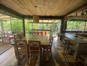 A wooden dining area with a large table and chairs, a bar with stools, and a view of greenery through large windows. - Sítio São Pedro da Serra (Nova Friburgo)