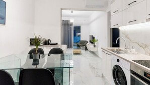 A modern kitchen with a washing machine, a glass dining table, and a view into a living area. - Sunny Nest Apartment 2 in Ixia (Ialisos)