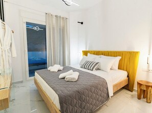 Quarto com cama, mesa de cabeceira de madeira, porta de vidro para uma varanda e um roupão branco pendurado em um gancho.