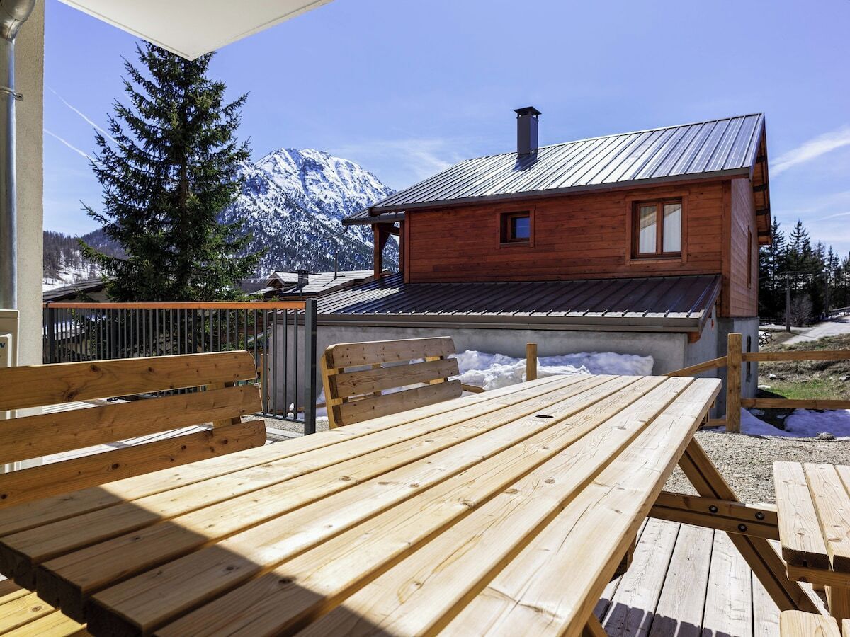 A wooden table and benches outside a cabin with a metal roof, surrounded by snow and mountains.