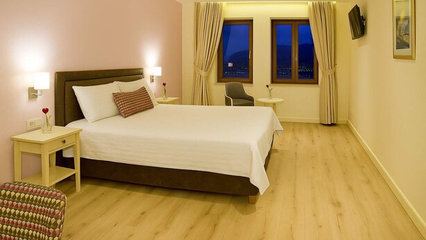 A hotel room with a large bed, bedside tables, a chair, a small table, and a window with curtains. - Arktos Hotel Superior Room w/ Stunning Lake View (Ioannina)