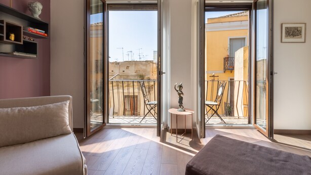 Un salon moderne avec un canapé, une petite table et une statue. La pièce est dotée de grandes portes vitrées qui donnent sur un balcon extérieur, aménagé avec des chaises et une table.
