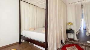 Un lit à baldaquin avec des rideaux blancs, un tapis rouge à motifs et une petite table de chevet avec une lampe.