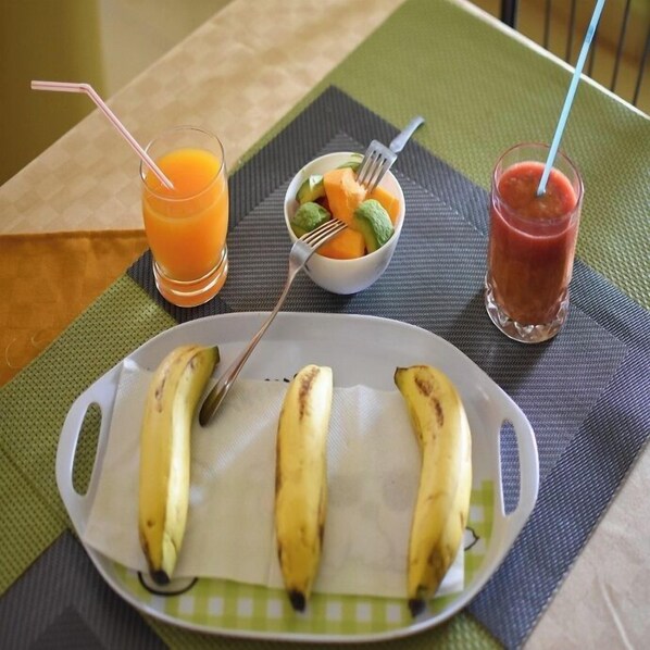 Un plato con plátanos, un bol de fruta y dos bebidas sobre una mesa.