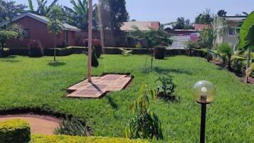 A well-maintained garden with a paved area, surrounded by lush greenery and a few buildings in the background.