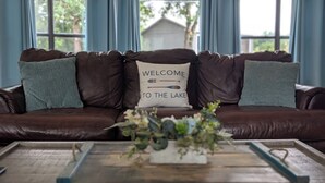 A brown leather sofa with a pillow that reads 'WELCOME TO THE LAKE' and a small plant on a wooden coffee table.