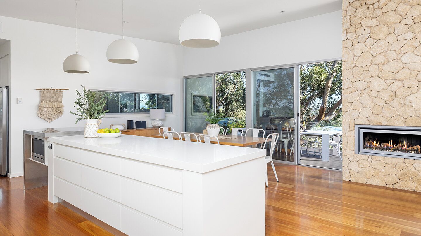 A modern kitchen with a large island, white cabinetry, pendant lighting, and a stone fireplace.