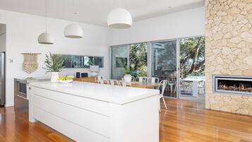 A modern kitchen with a large island, white cabinetry, pendant lighting, and a stone fireplace.