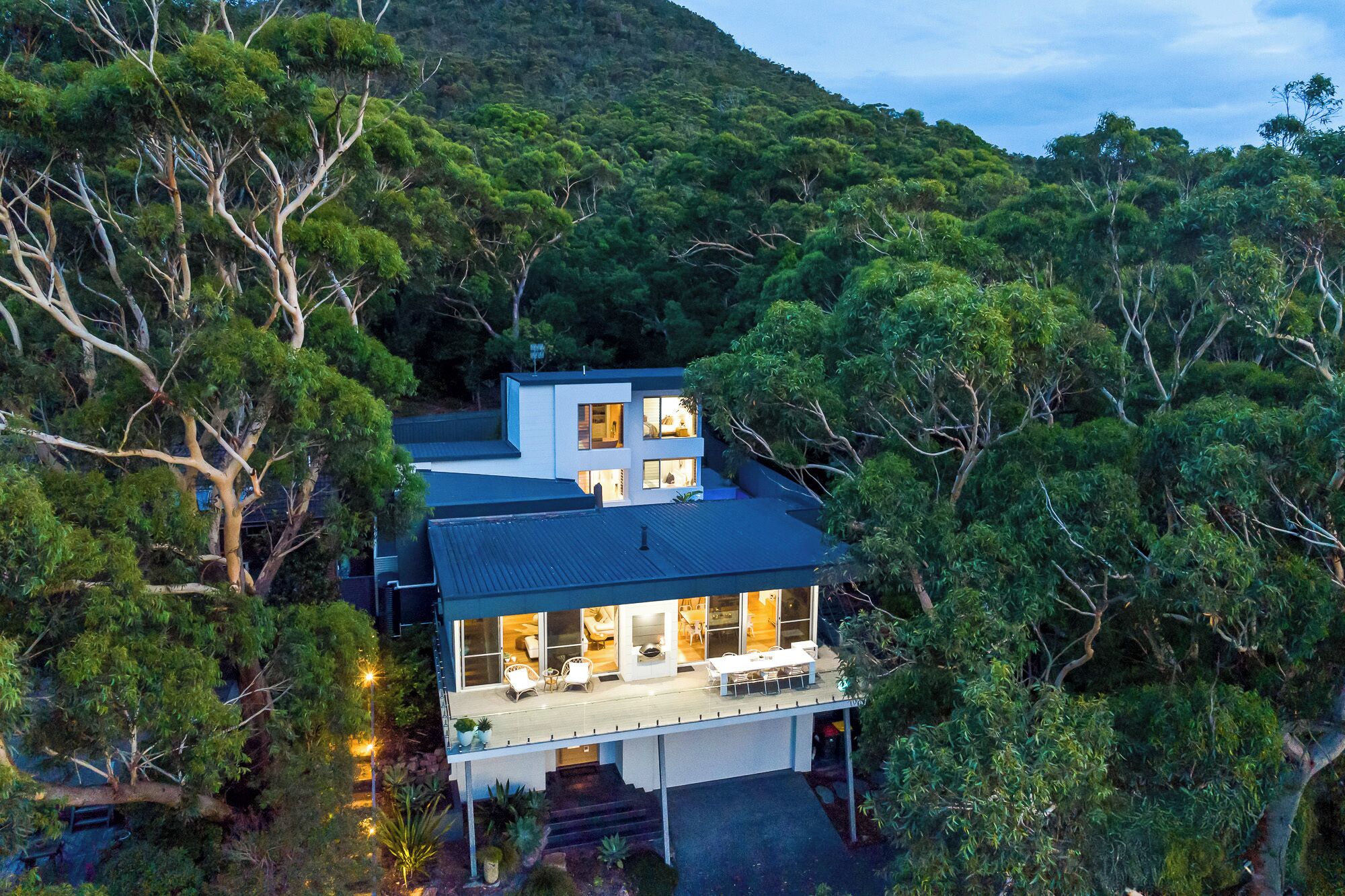 A modern two-story house surrounded by dense forest, with a large balcony and multiple windows.