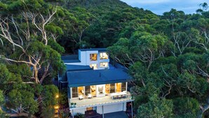 Una casa moderna de dos pisos rodeada de un bosque frondoso, con un balcón grande y varias ventanas.
