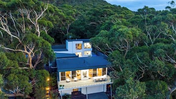 A modern two-story house surrounded by dense forest, with a large balcony and multiple windows.