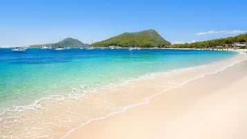 A beach with clear turquoise water, boats, and a mountain in the background.