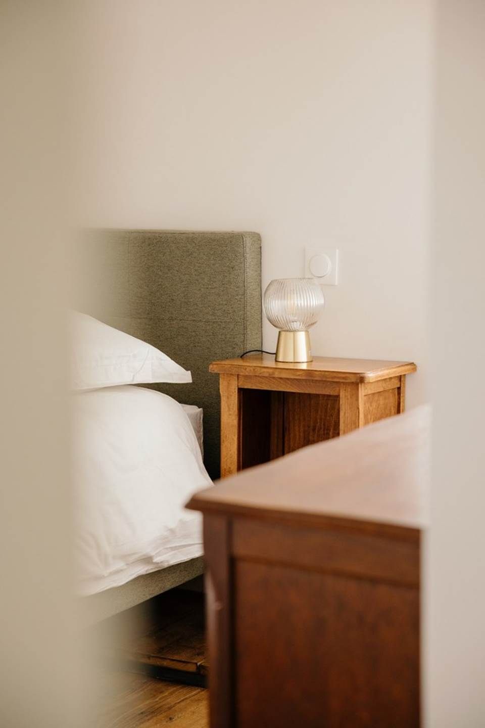 A neatly made bed with white linens, a wooden nightstand with a lamp, and a wall switch.
