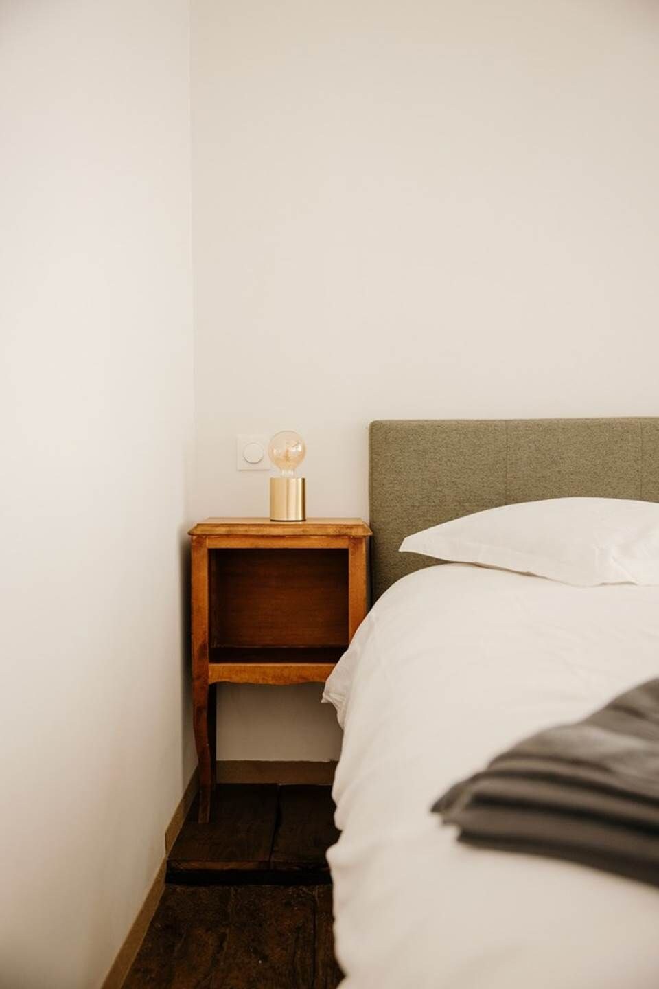 Un dormitorio en esquina con mesita de noche de madera, lámpara de noche, cama con ropa blanca y cabecero gris.
