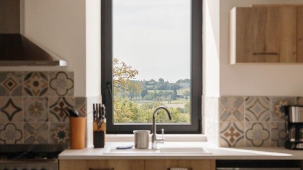 Comedor con mesa dispuesta para cuatro personas, ventana con vista a vegetación y cocina con armarios de madera y estufa.