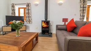 A living room with a wooden coffee table, a sofa with red cushions, a TV, and a wood-burning stove. - Woodland Roost (Bideford)