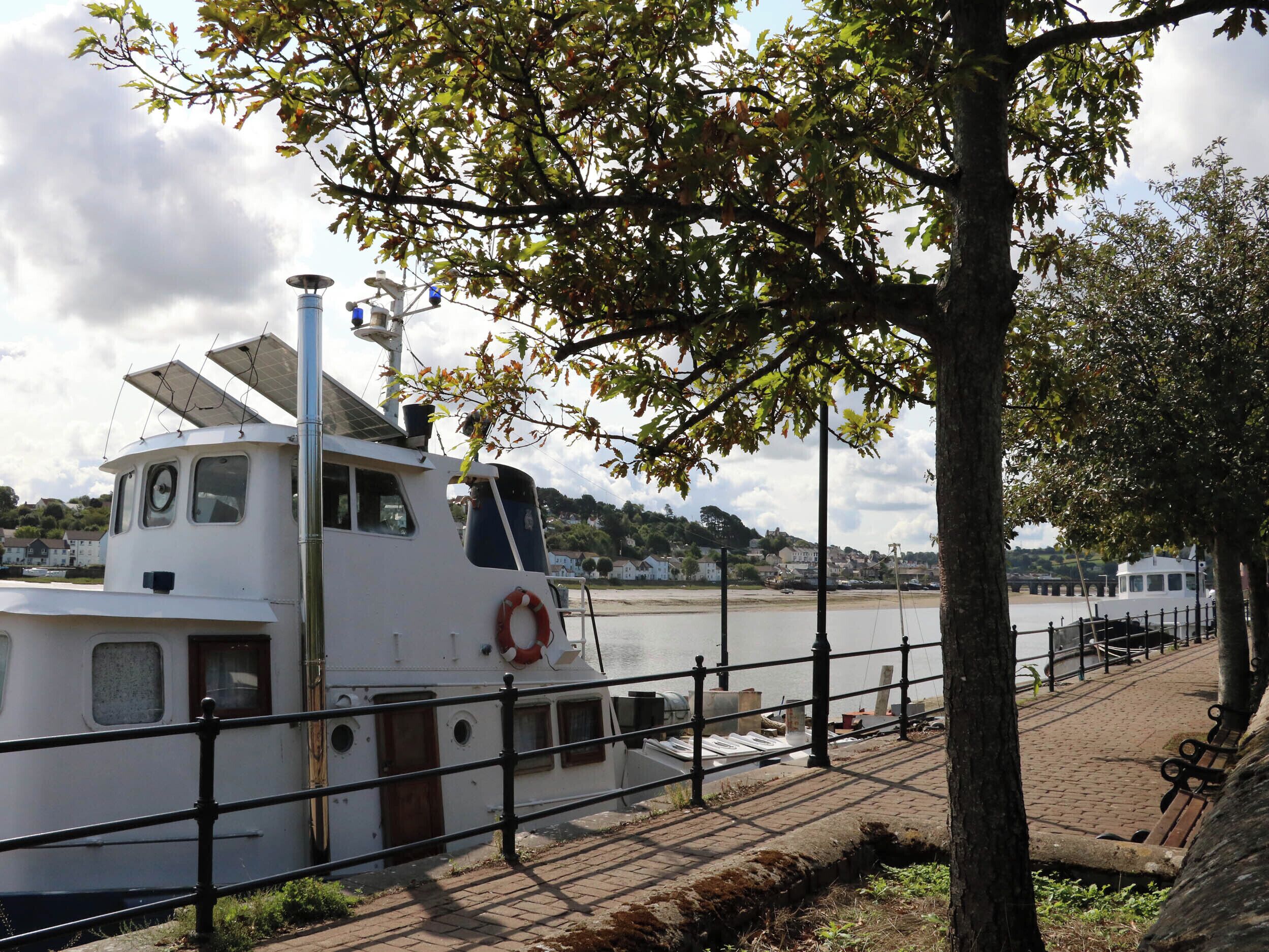A white boat docked at a pier with a lifebuoy attached to the side, a brick walkway, and a tree in the foreground.