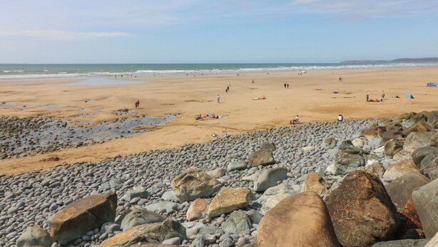 Une plage avec des galets et des rochers au premier plan, une plage de sable au milieu, et l’océan à l’arrière-plan.