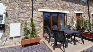 Un bâtiment en pierre doté d’un patio comprenant une table et des chaises, un jardinier en bois avec des plantes et une enseigne indiquant le nom du bâtiment.