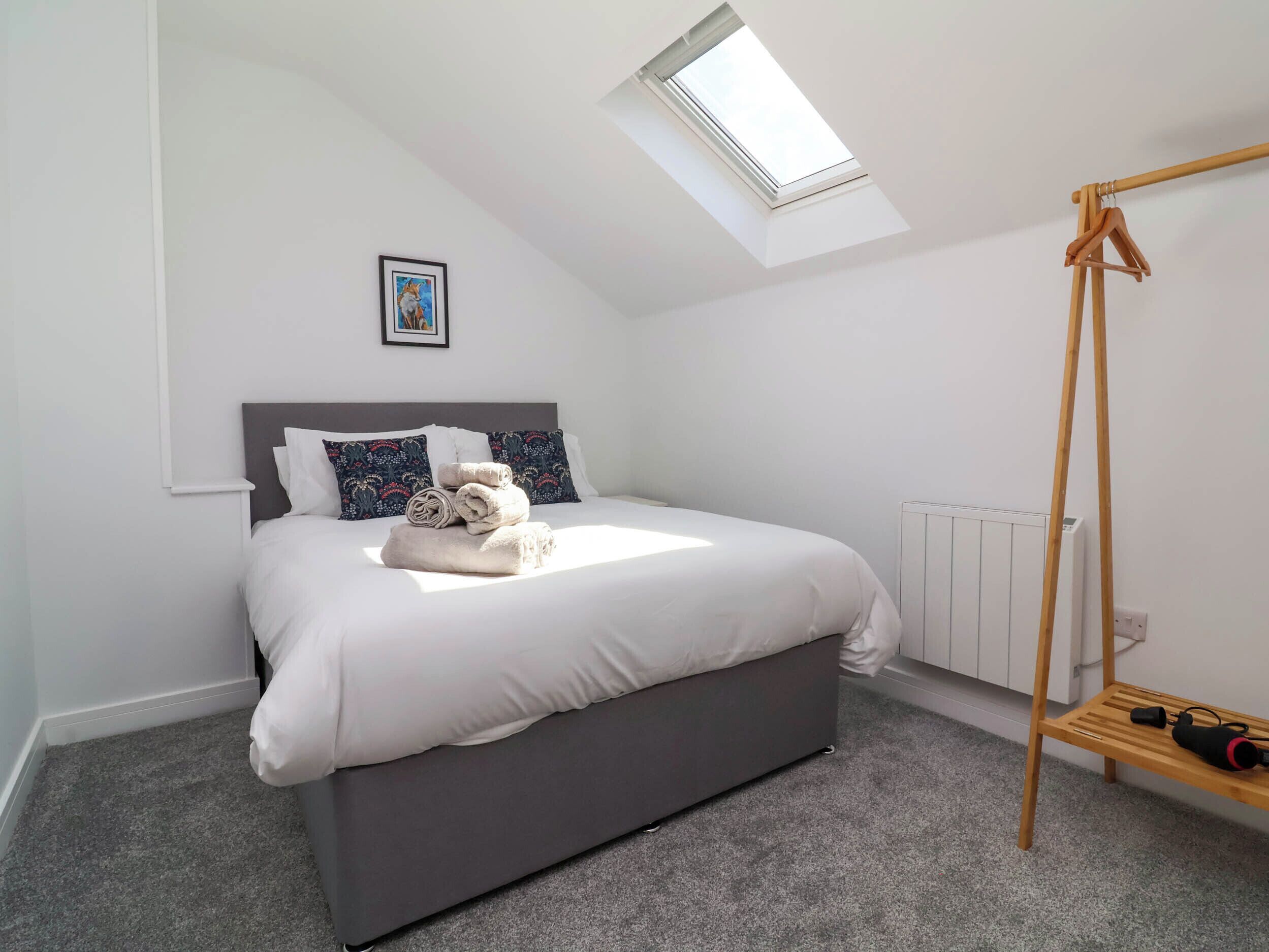 A modern bedroom with a skylight, a bed with white linens, a grey headboard, and a wooden drying rack.