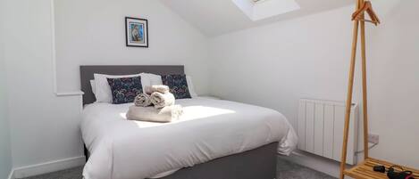 A modern bedroom with a skylight, a bed with white linens, a grey headboard, and a wooden drying rack.