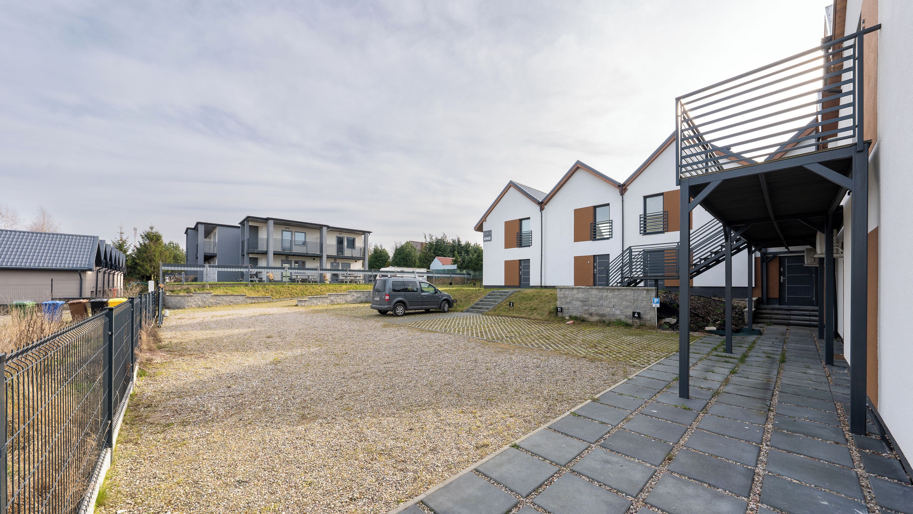 Et moderne boligområde med grusvej, en bil og en bygning med trappe og balkon.
