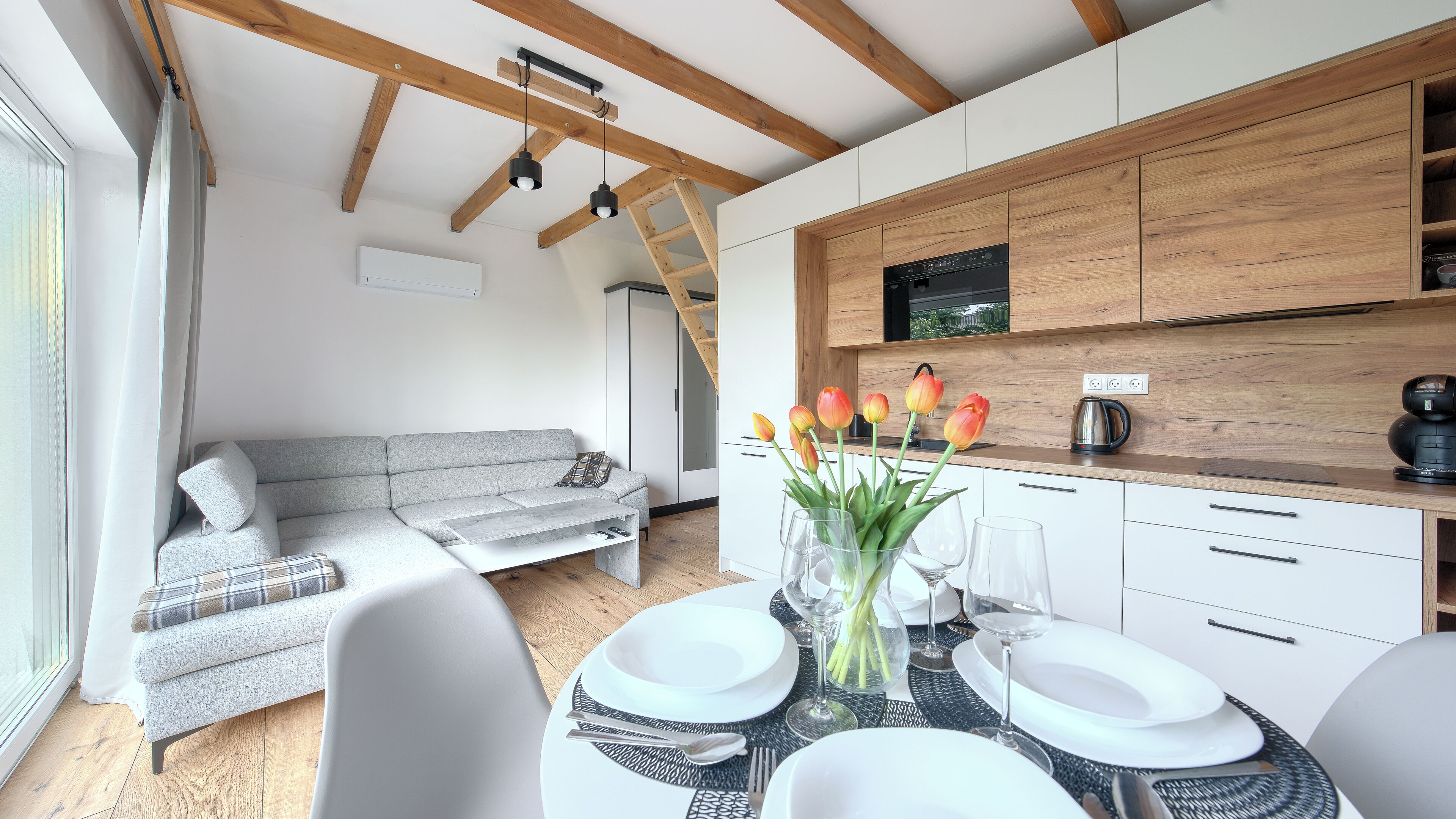 A modern kitchen with wooden cabinets, a dining area with a table set for two, and a grey sofa in the living area.