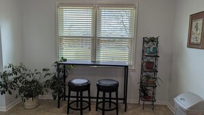 A room with a window, two stools, a table, a potted plant, a metal plant stand, and a trash bin. - Moontown Magic (Huntsville)