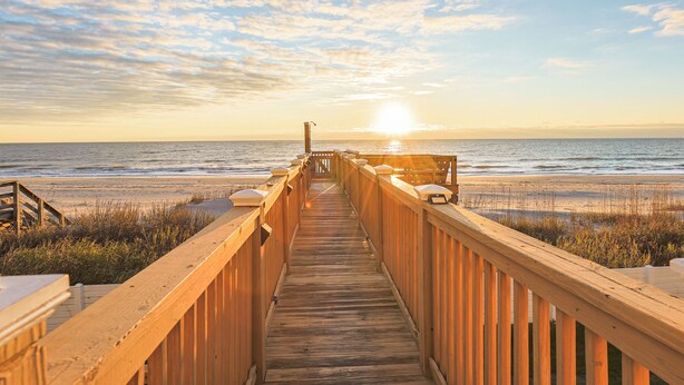 Une promenade en bois menant à une plage, avec un coucher de soleil en arrière-plan.