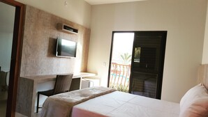 Een hotelkamer met een bed, een aan de muur bevestigde televisie, een klein bureau en een balkon met uitzicht op palmbomen.