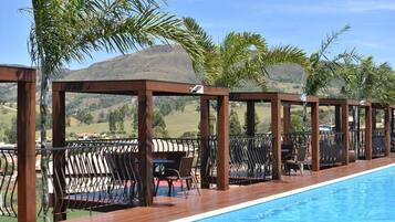 A poolside area with wooden pergolas, lounge chairs, and tables, overlooking a mountainous landscape.