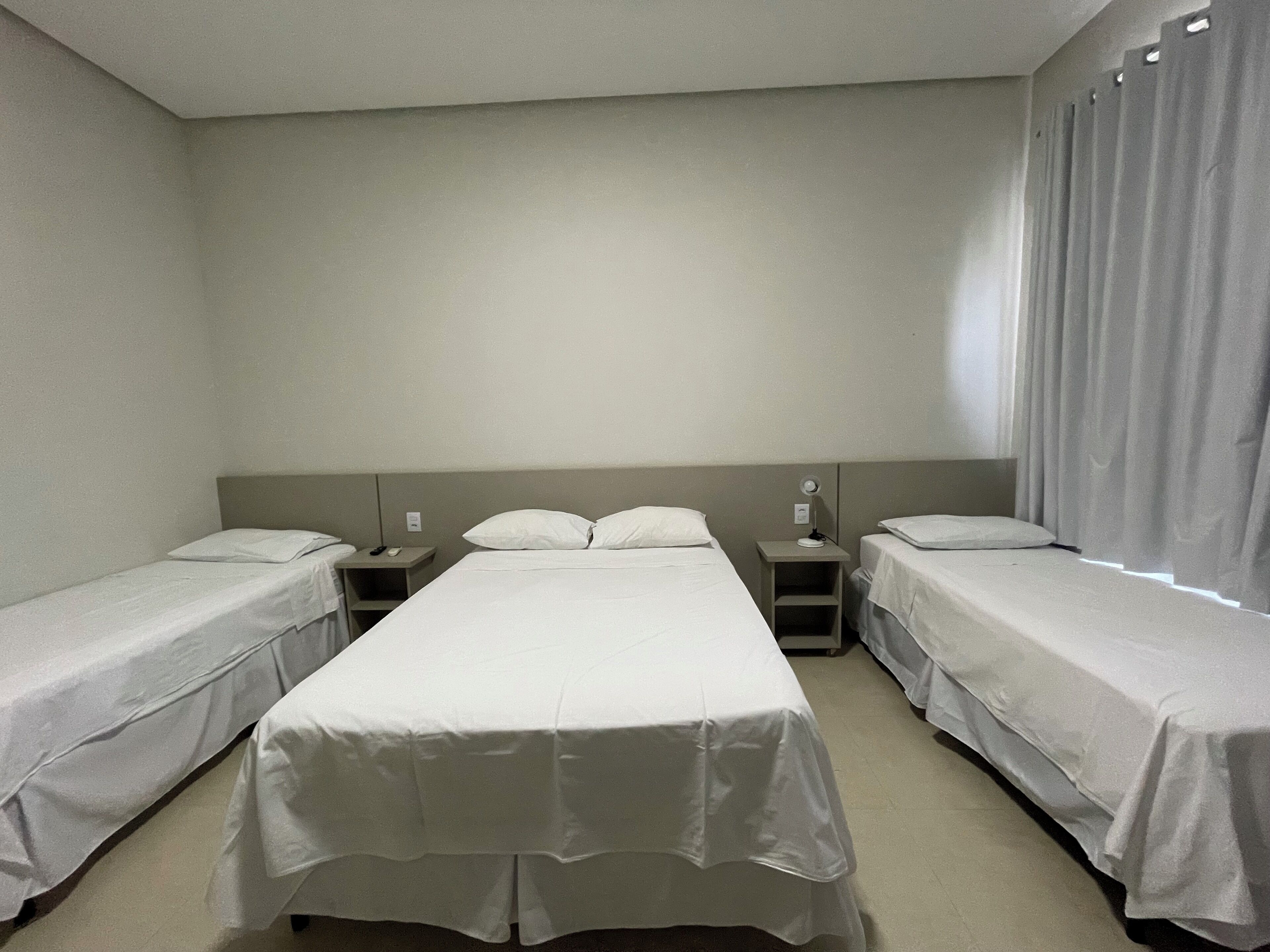 Ein Hotelzimmer mit drei Einzelbetten, einem kleinen Tisch und einem Nachttisch mit Lampe.