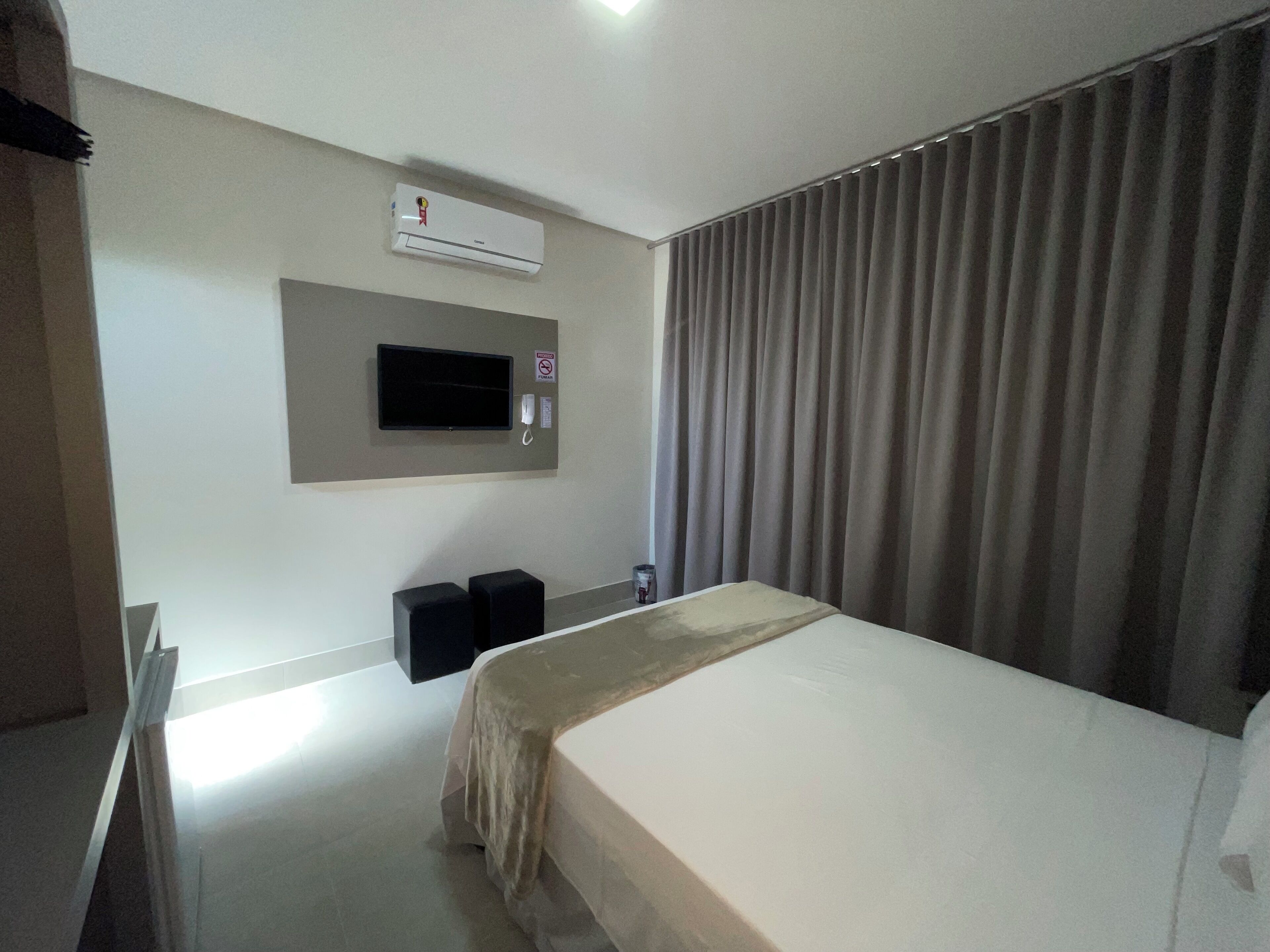 A hotel room with a bed, a TV mounted on the wall, an air conditioning unit, and a curtain.