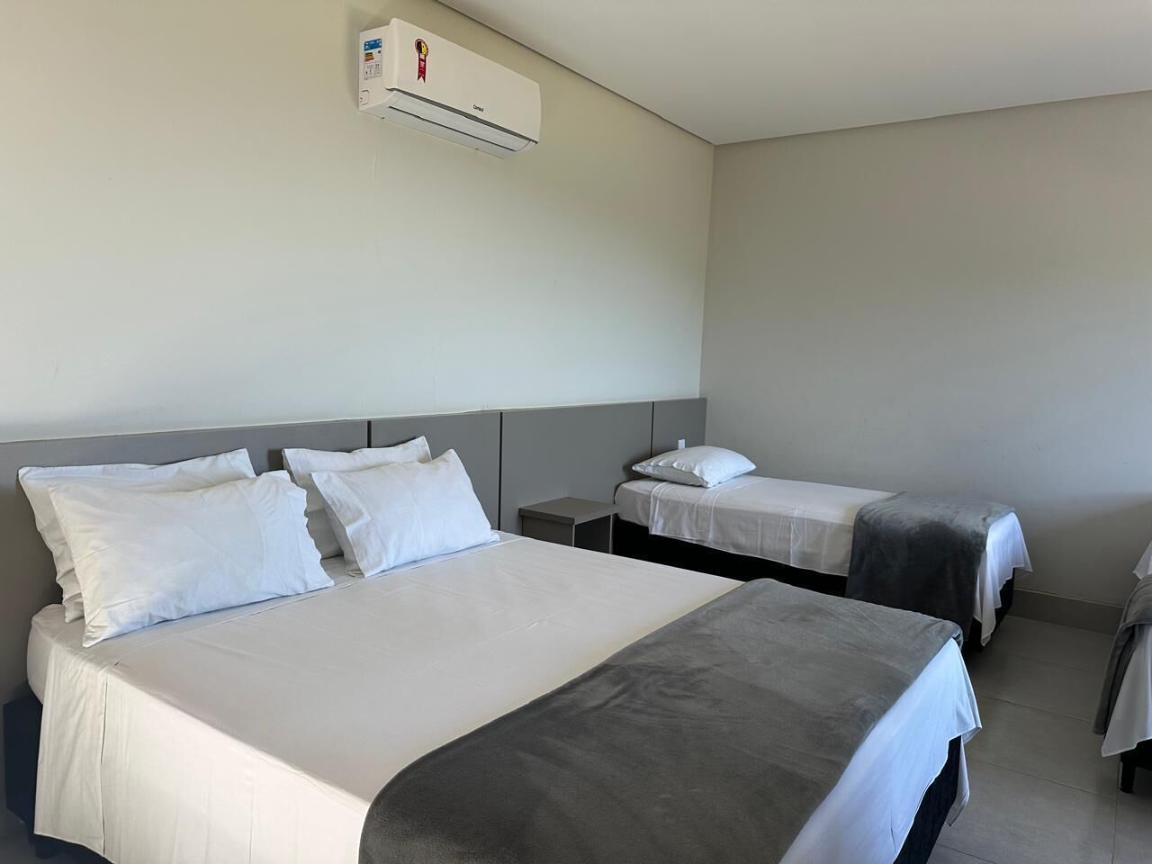 Habitación de hotel con una cama grande, dos camas más pequeñas, una mesita de noche y un equipo de aire acondicionado en la pared.