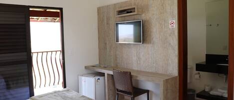 Habitación de hotel con cama, escritorio con televisor, refrigerador pequeño, una silla y baño visible a través de una puerta abierta.
