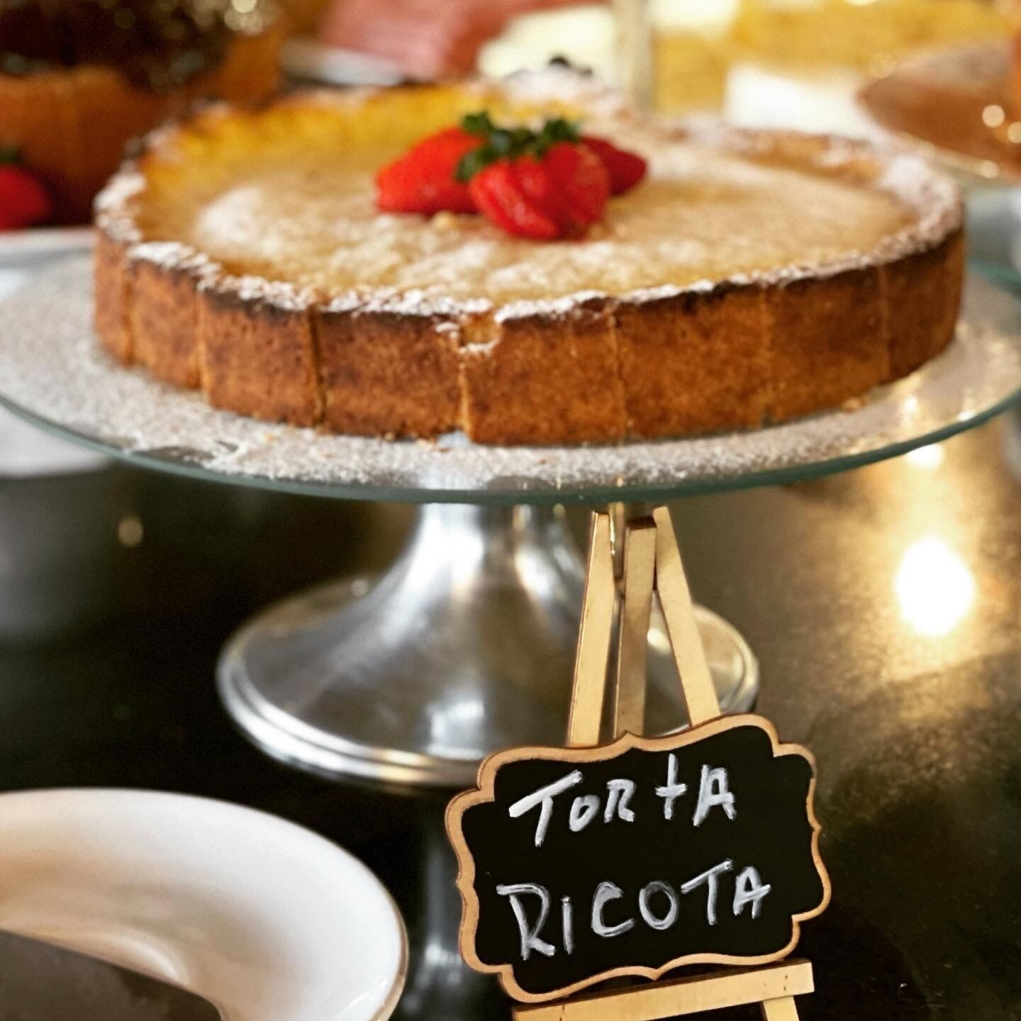 A cake on a glass stand with a sign that reads "Torta Ricota".
