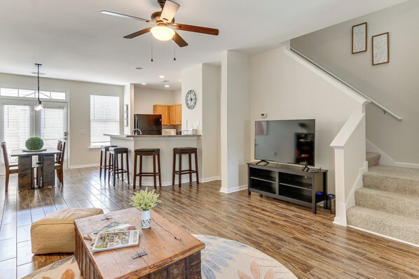 A spacious living room with hardwood flooring, a ceiling fan, a dining area with a table and chairs, a kitchen area with a refrigerator and microwave, and a staircase.