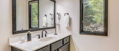 Un baño con un espejo grande, un lavabo, un perchero para toallas y una ventana con vistas a árboles.